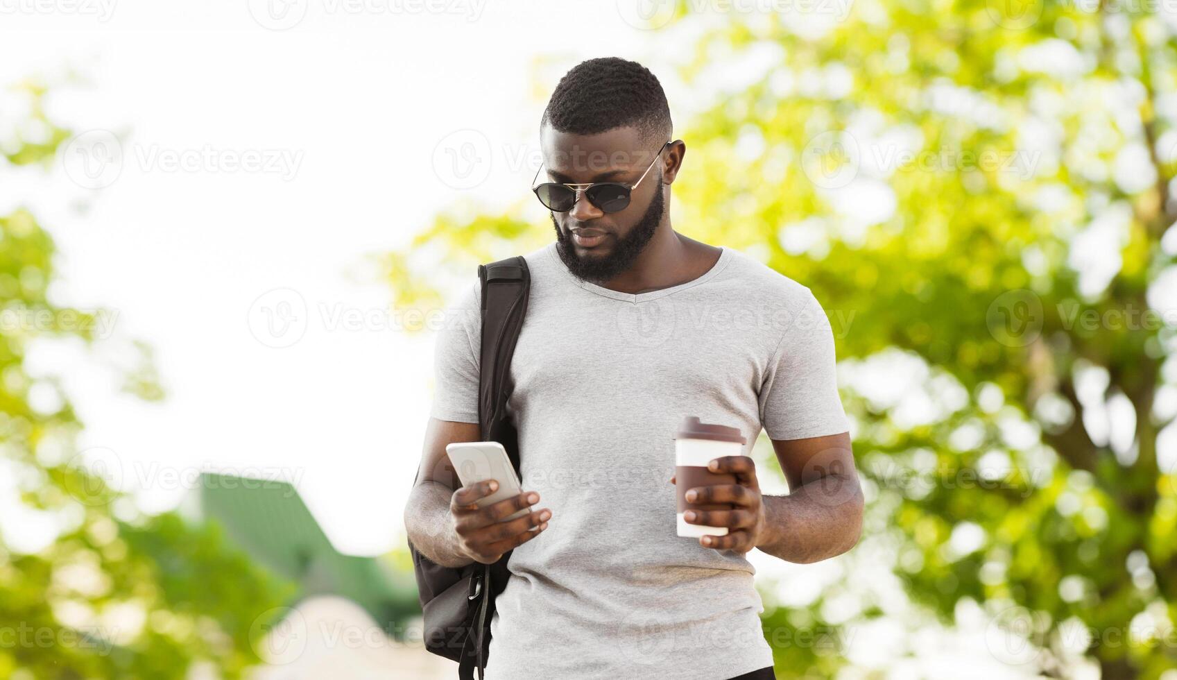 A man in sunglasses, wearing a grey t-shirt, uses his phone while holding a cup of coffee. photo