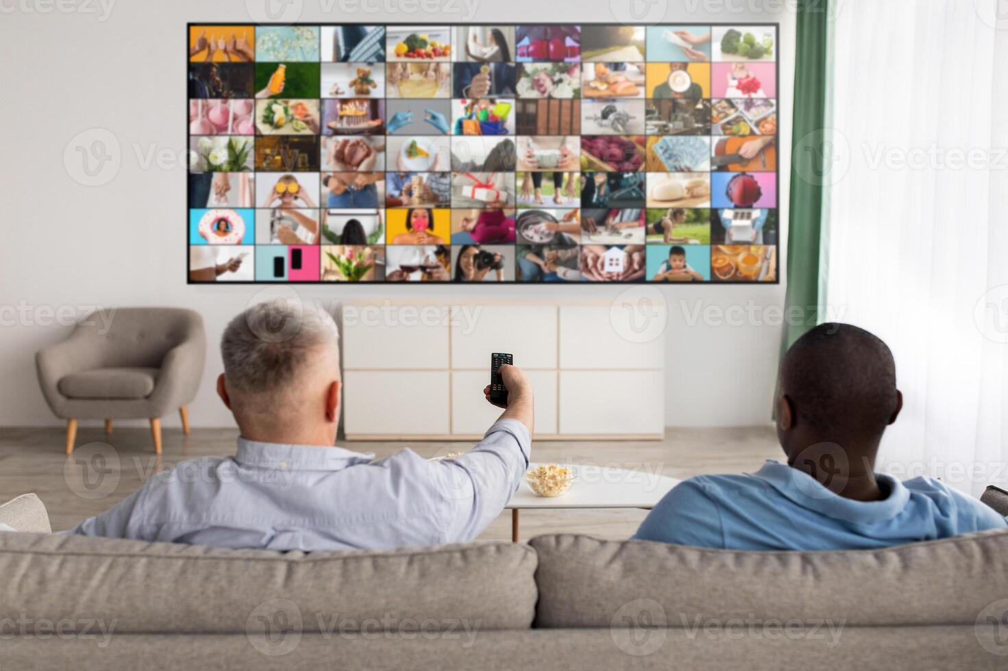 Two men are sitting on a couch in a living room, watching a large TV screen displaying a grid of various images. The man in the foreground is holding a remote control and is looking at the TV. photo
