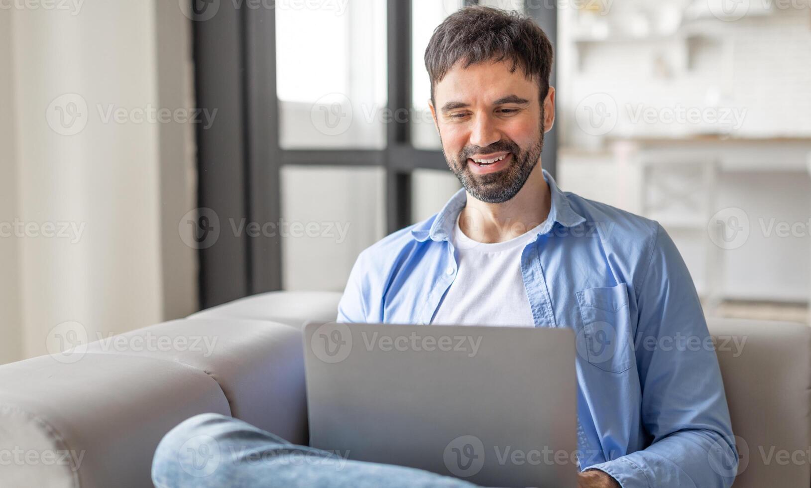 A man is sitting on a couch at home, using his laptop. He is smiling and appears to be enjoying his time online. The man is wearing a blue shirt and jeans, and the couch is white. photo