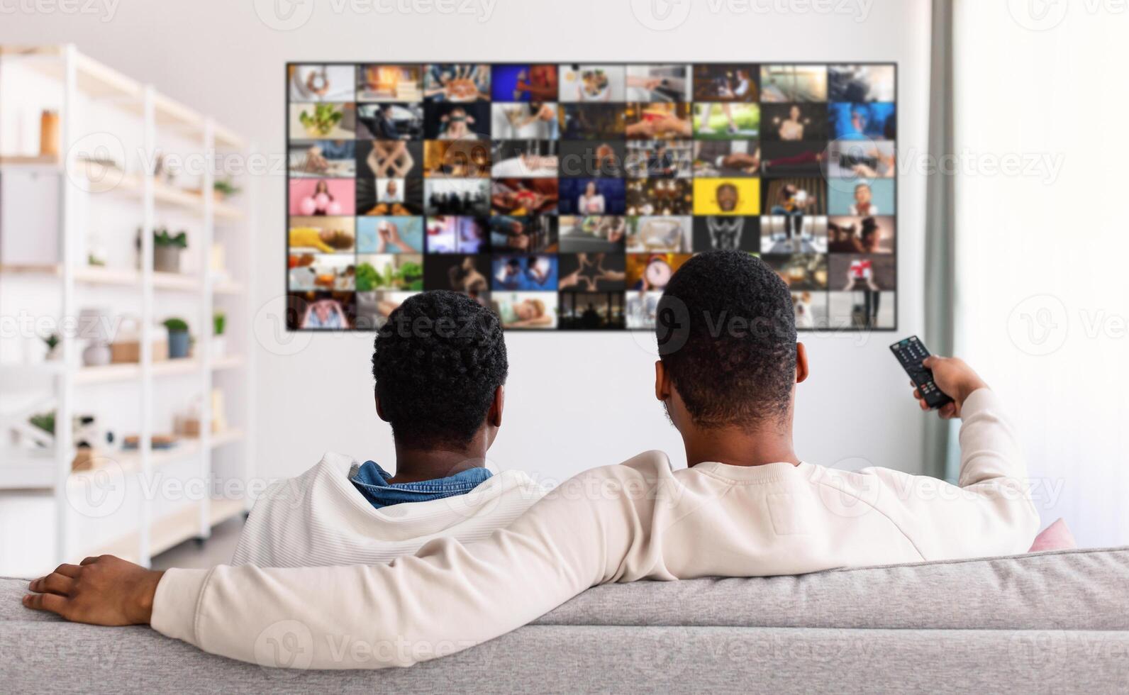Two men sit on a gray couch watching television in a living room. The man on the left is wearing a white hoodie and the man on the right is holding a remote control photo