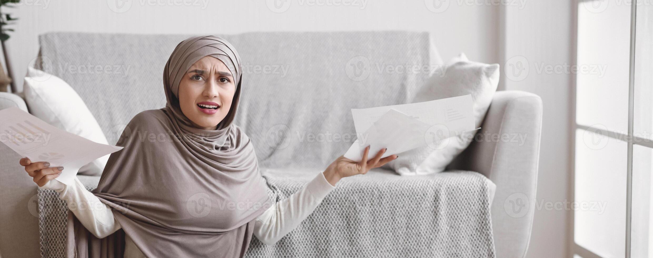 My Brain Is Full. Concerned Muslim Girl Freelancer Holding Papers With Grumpy Face Expression While Working On Laptop On Floor At Home photo