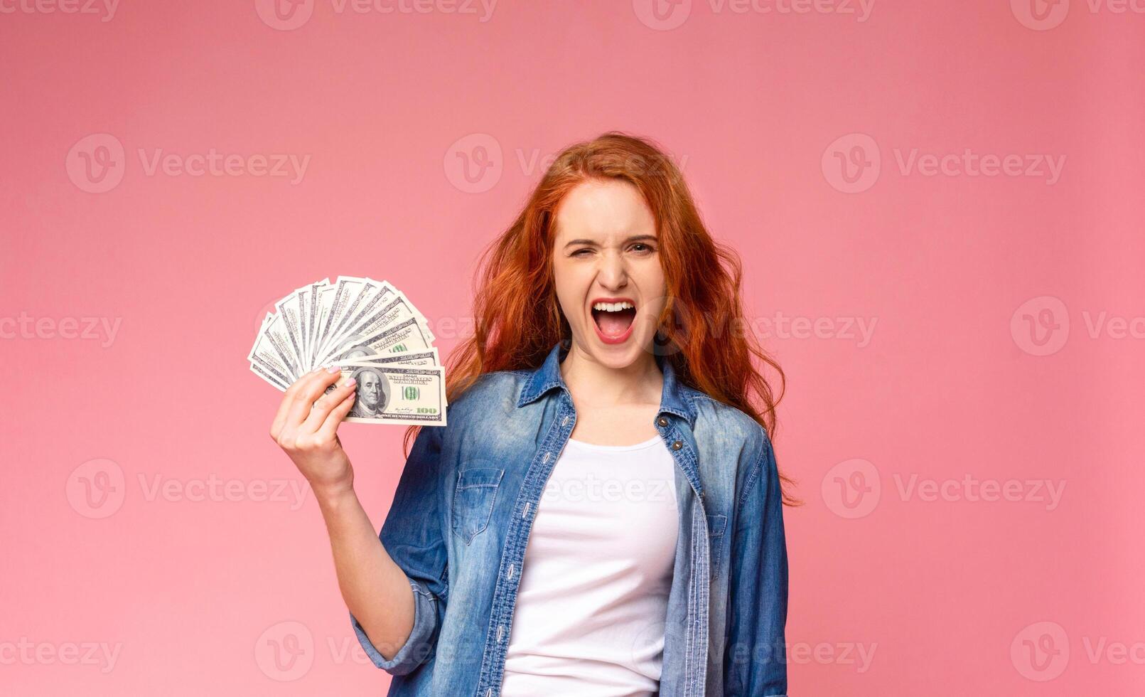 A woman with vibrant red hair is seen in the image, holding a fan of money in her hand. She appears to be posing confidently, showcasing the stack of bills photo