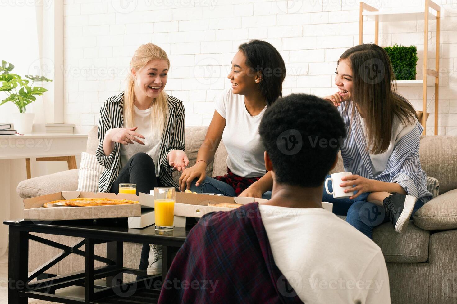 A small group of multiracial friends is seated around a coffee table in a well-lit living room, sharing laughter and stories over a meal of pizza and refreshing beverages. photo