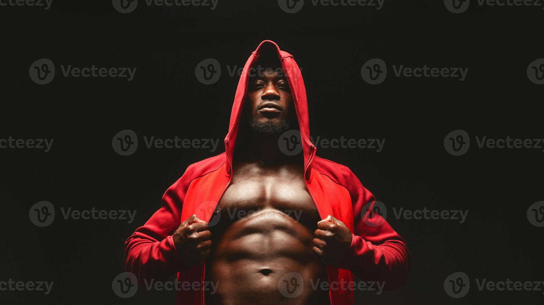 Confident african bodybuilder in red hooded shirt with bare chest, black studio background photo
