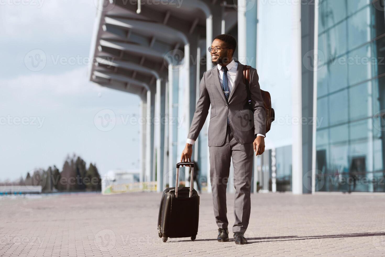 African American businessman is walking with his luggage outside a modern airport. The mood is dynamic and professional, highlighting travel and business with a sleek airport backdrop. photo