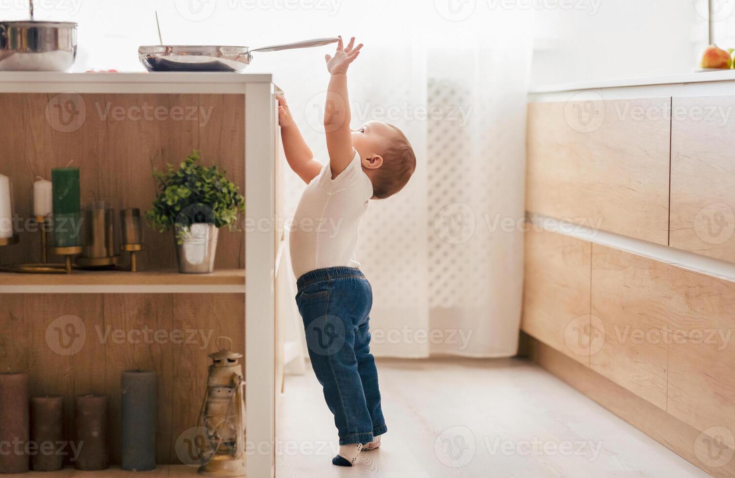 A curious toddler stands on tiptoe in a modern kitchen, reaching for a frying pan on the counter, illuminated by morning sunlight. photo