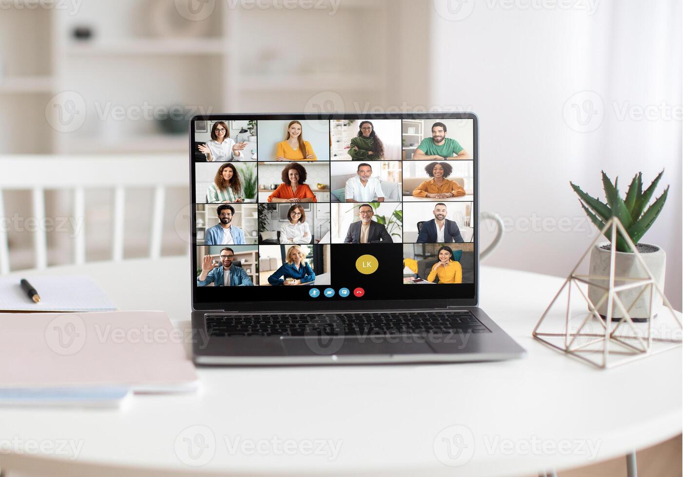 A laptop on a white table displays a virtual team meeting with multiple participants. The bright, modern home office background includes a white bookshelf, green plant, and geometric decor piece. photo