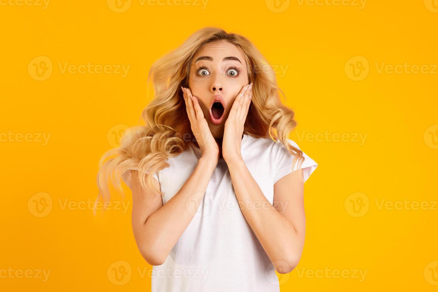 This photo shows a young woman with blonde hair and a white t-shirt standing in front of a yellow background. She is looking at the camera with a surprised expression