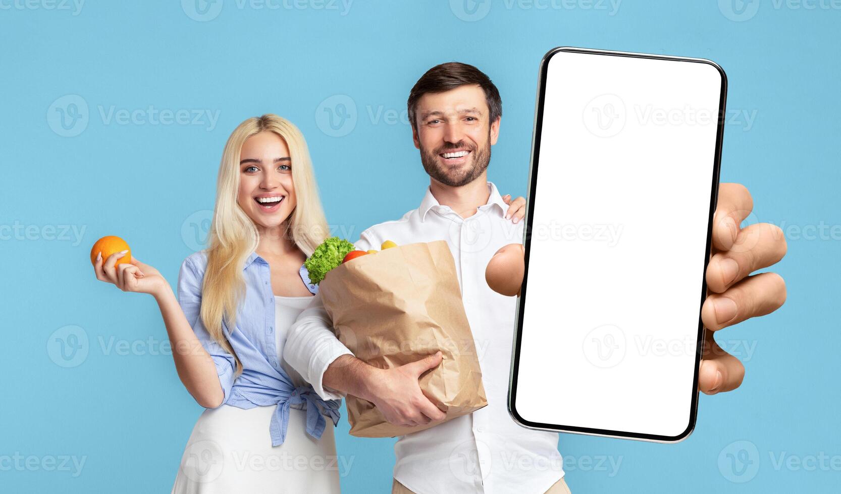 A man and woman stand side by side, both smiling at the camera. The woman is holding an orange and the man is holding a paper bag full of groceries and a smartphone with a blank white screen photo