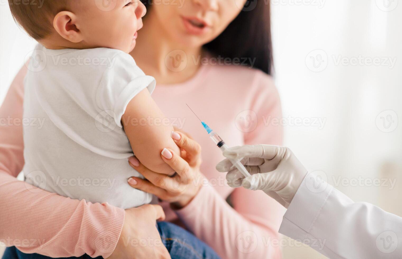 A mother comforts her crying baby as a doctor prepares to administer a vaccine. The infant is held in a secure embrace, showcasing maternal care during a routine immunization procedure. photo