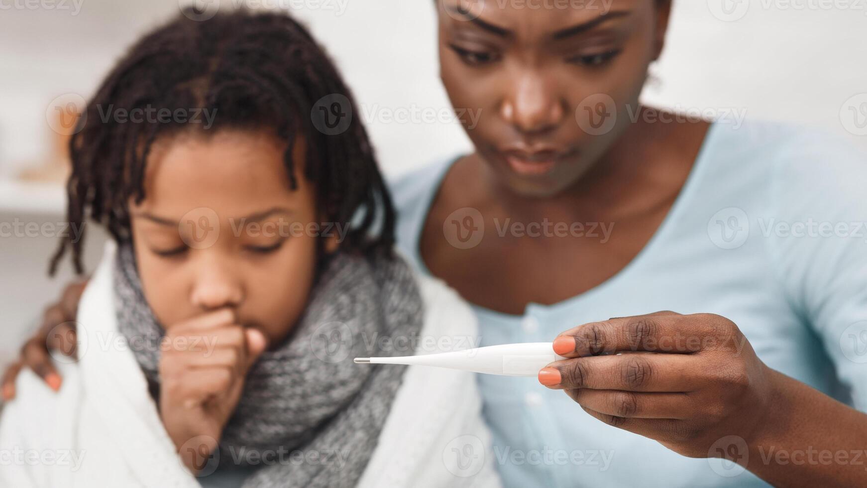 Cold Symptoms. Worried black mother looking at thermometer, ill daughter coughing. Selective focus photo