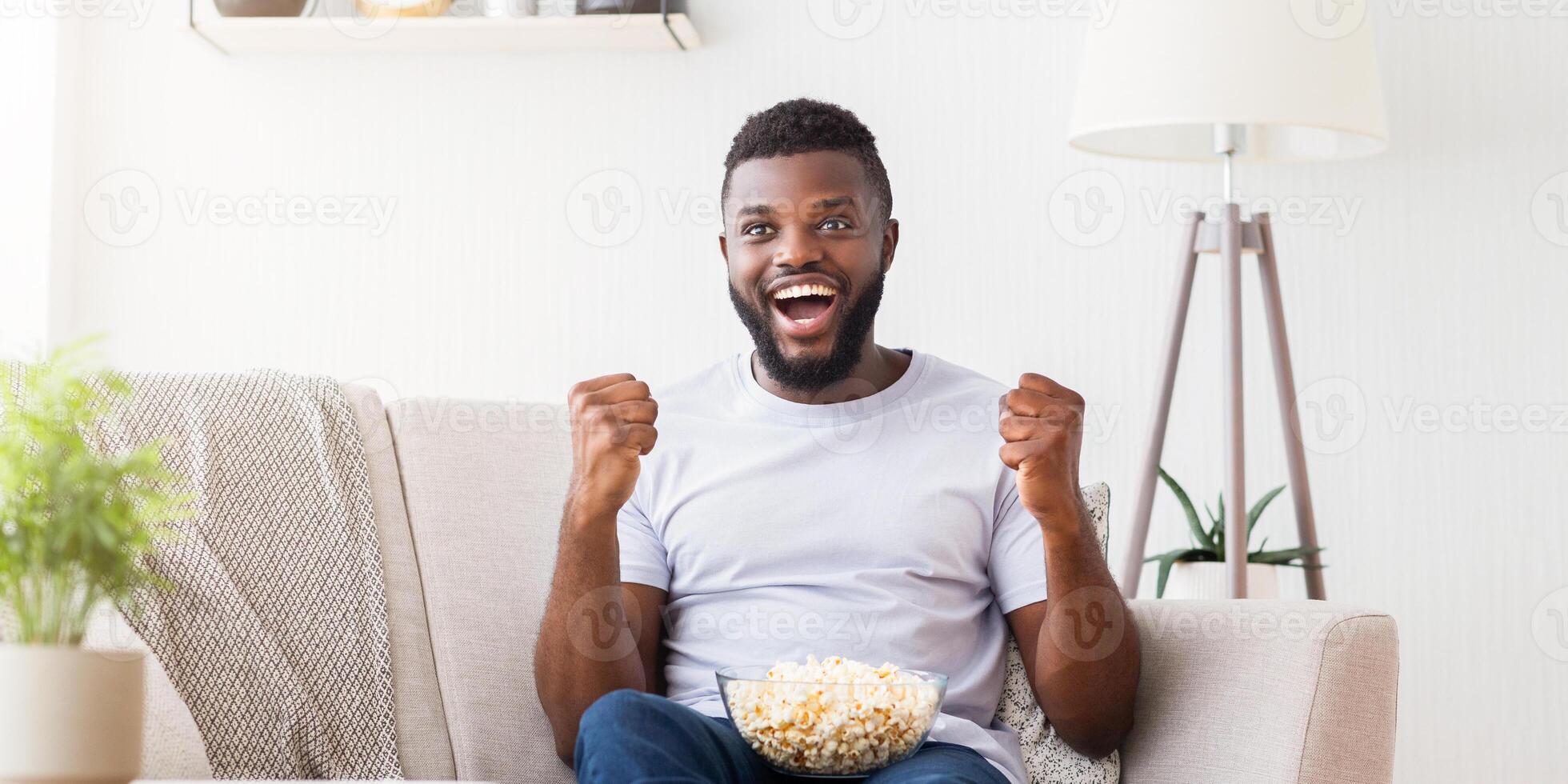 Black man sits on a white couch, his arms raised in excitement, while he watches television. He is wearing a white t-shirt, blue jeans, and has a bowl of popcorn in his lap photo