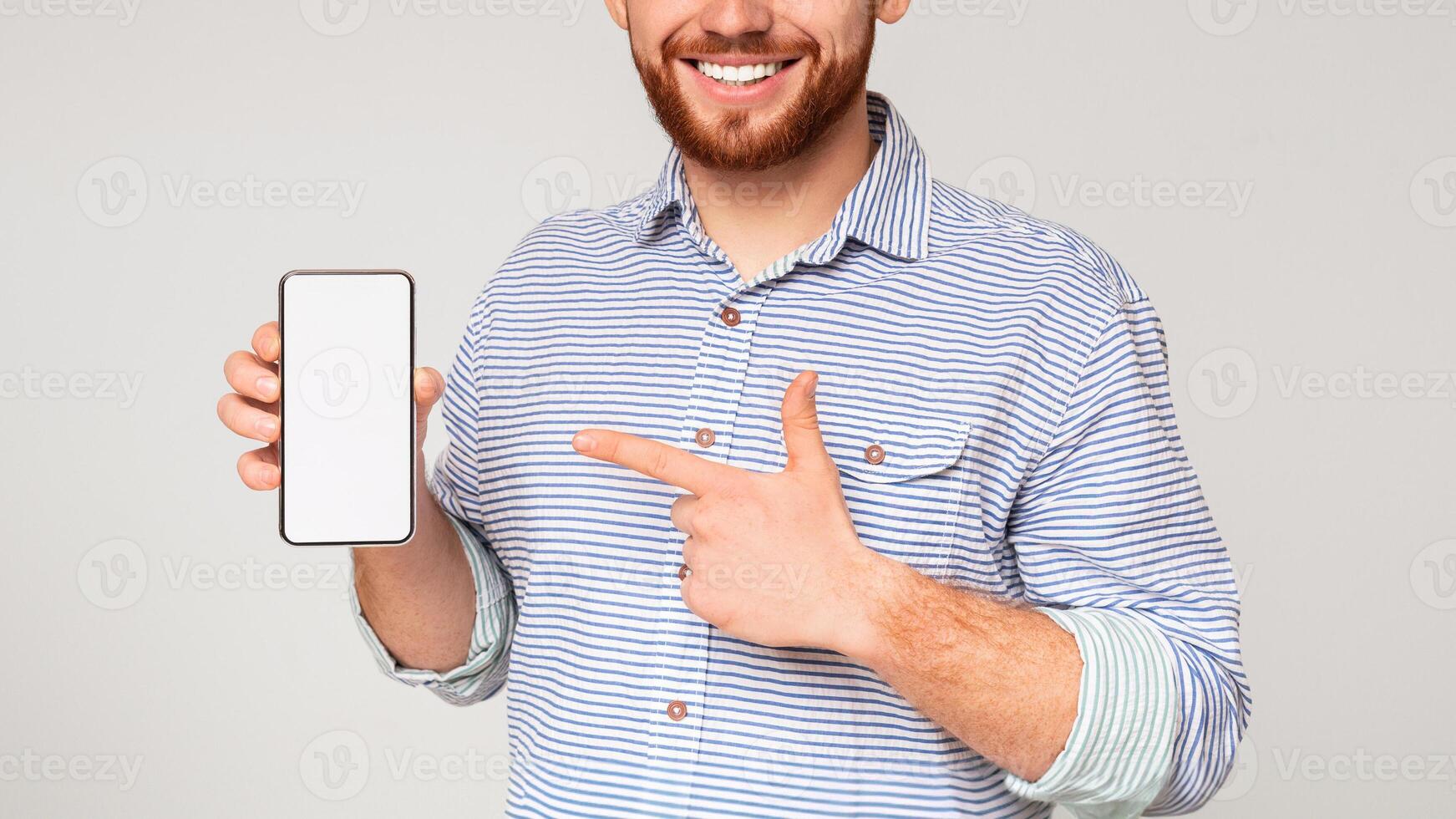Smiling millennial man holding latest slim cellphone with blanc screen and pointing at it, panorama, free space photo