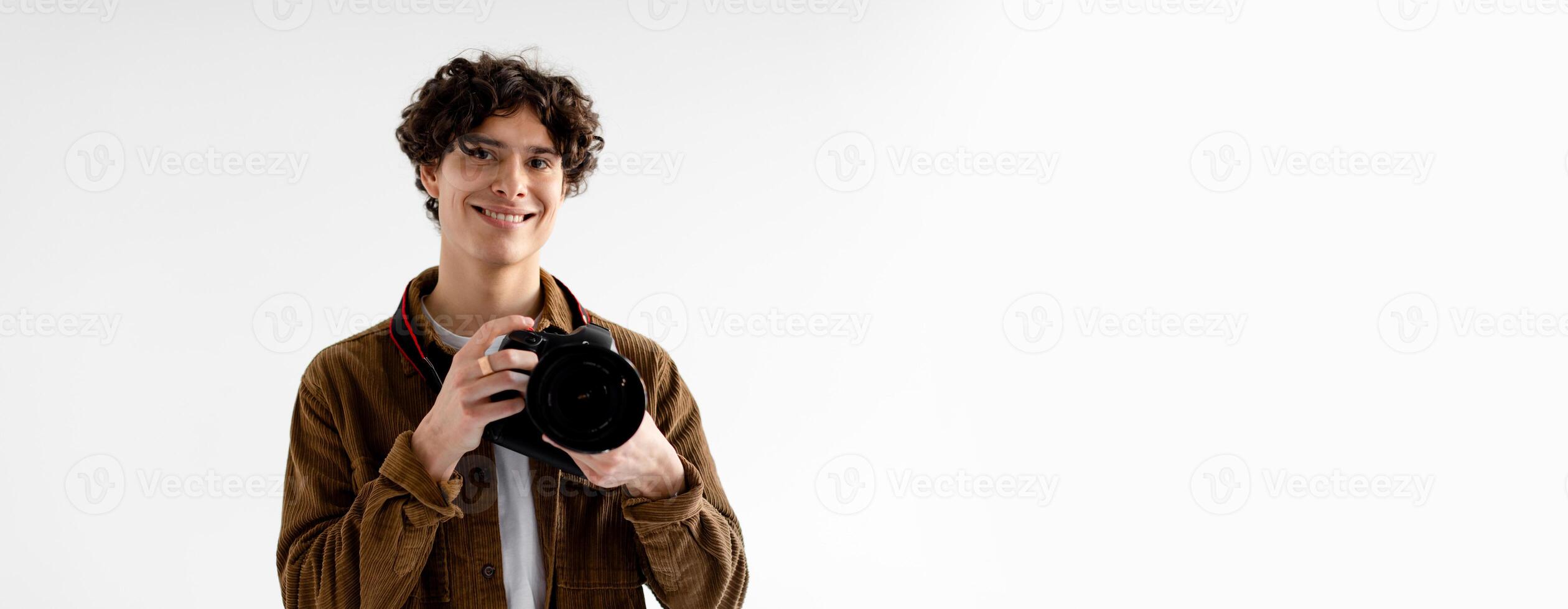 A young man with curly hair stands in a bright studio, holding a camera and smiling. The backdrop is light-colored, providing a professional atmosphere for photography, copy space photo
