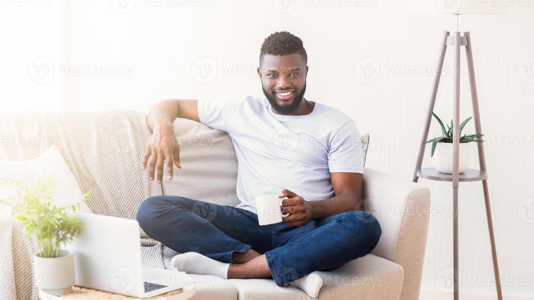 Black man in a white t-shirt and jeans sits comfortably on a couch in a living room. He is holding a white mug and smiling at the camera. There is a laptop on the coffee table in front of him photo
