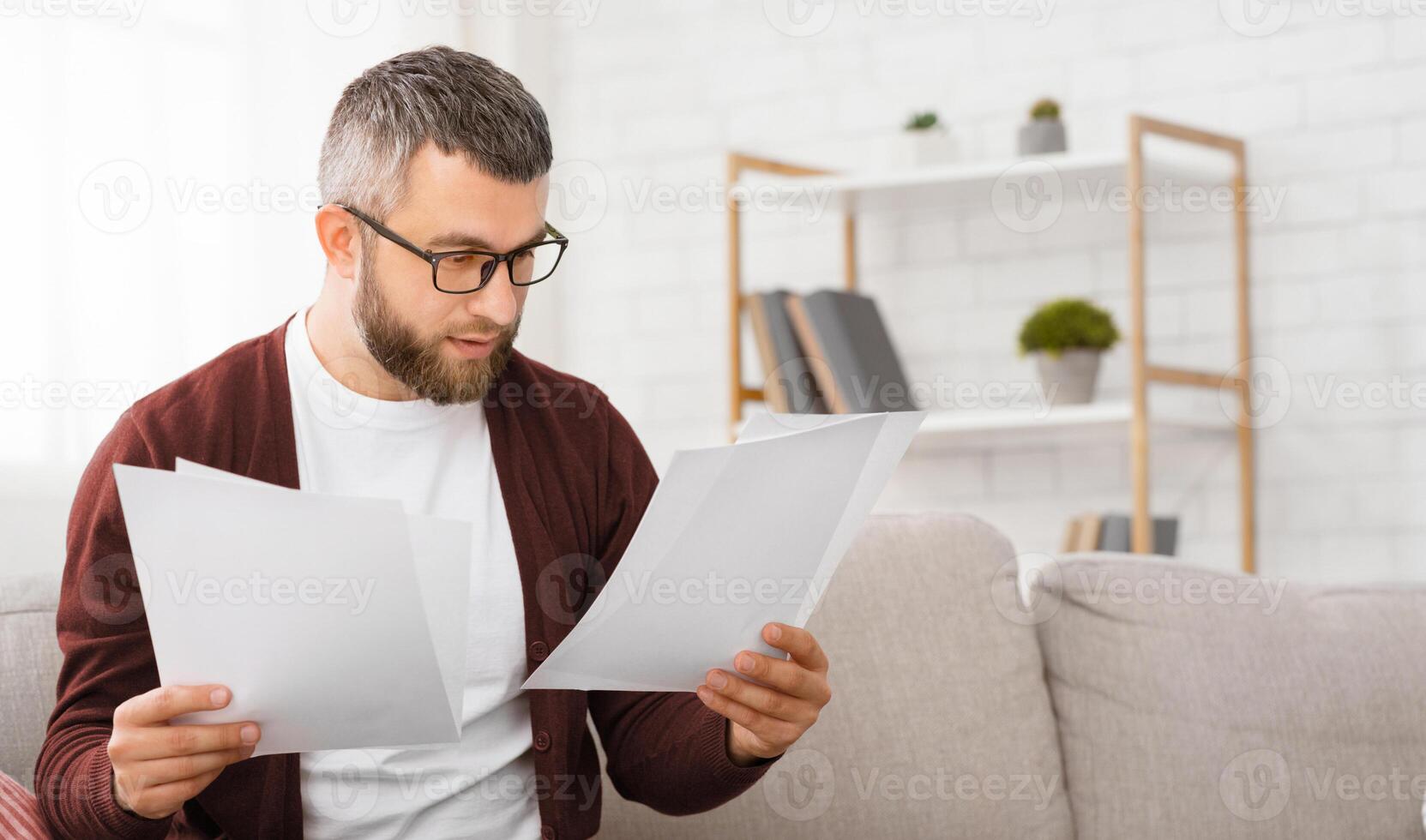 A man is seated on a couch, attentively reading a piece of paper in his hands. His concentration is evident as he scans through the document, absorbed in its contents. photo