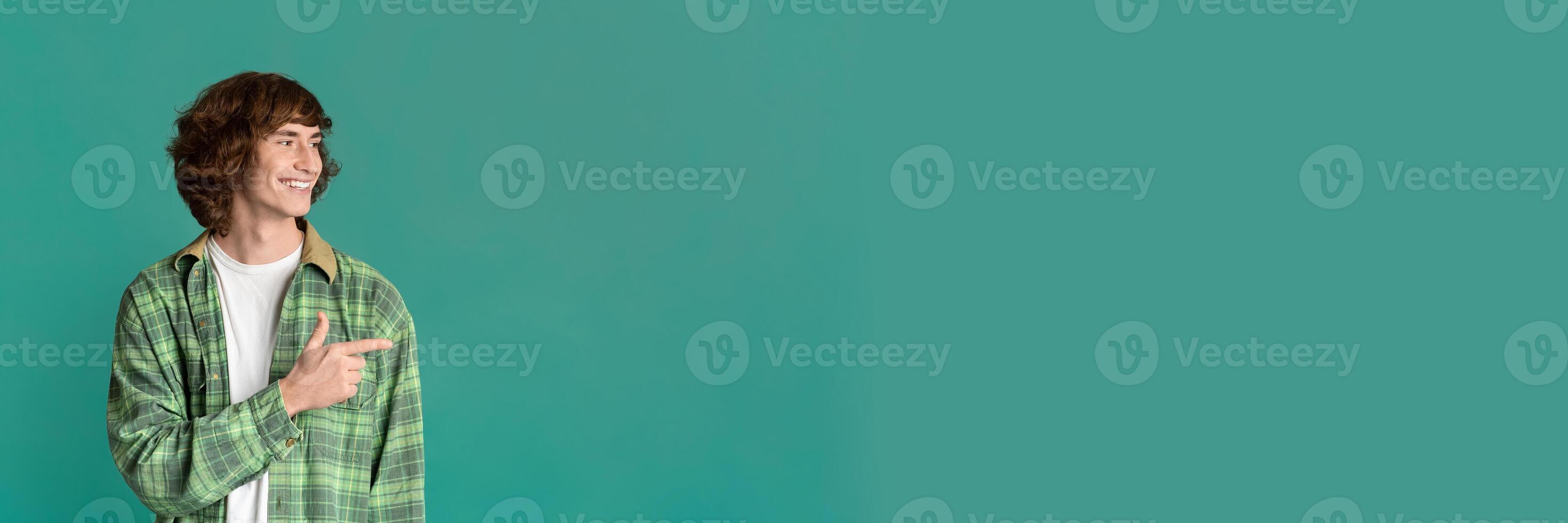 Cool stylish guy with curly hair pointing aside on color studio background, copy space for design, panorama photo