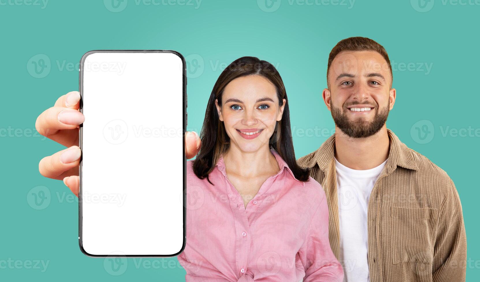 A couple, a woman in a pink shirt and a man in a tan corduroy jacket, stands in front of a teal background, smiling. The woman is holding a smartphone with a blank white screen, facing the camera. photo