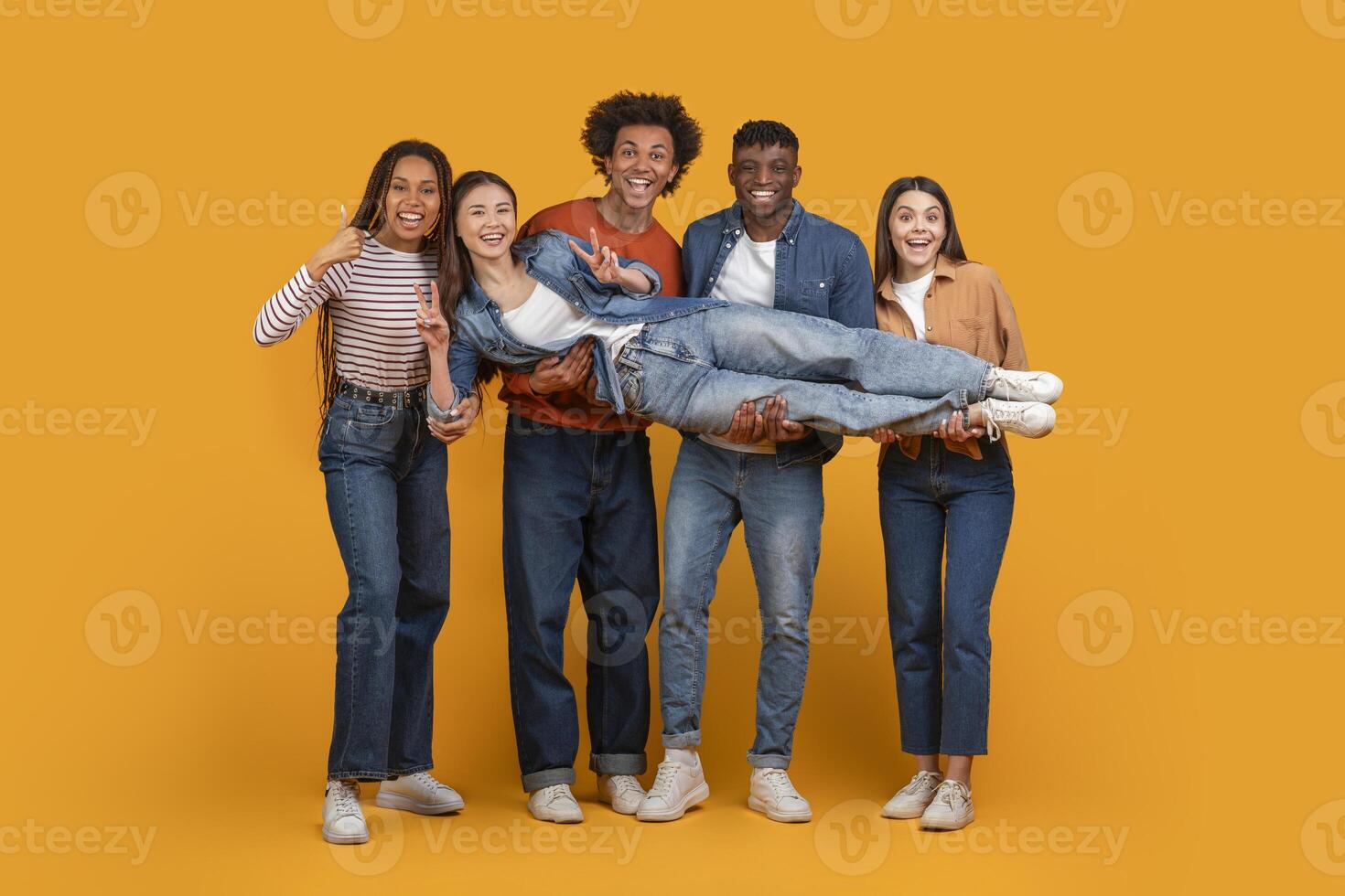 A group of four friends are holding up another friend in a studio setting. They are all smiling and appear to be having a fun time together. The background is a bright yellow. photo
