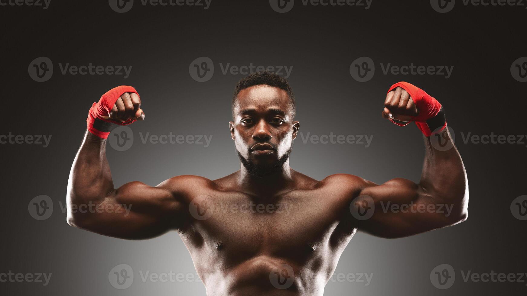 A shirtless man with red hand wraps flexes his bicep muscles in a studio setting. photo