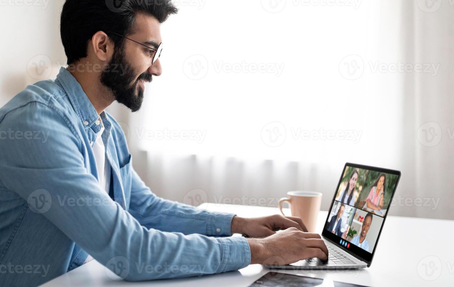 A young man is engaged in a virtual meeting using his laptop at his home office. He sits at a desk with a bright window in the background, interacting with multiple colleagues on the screen. photo