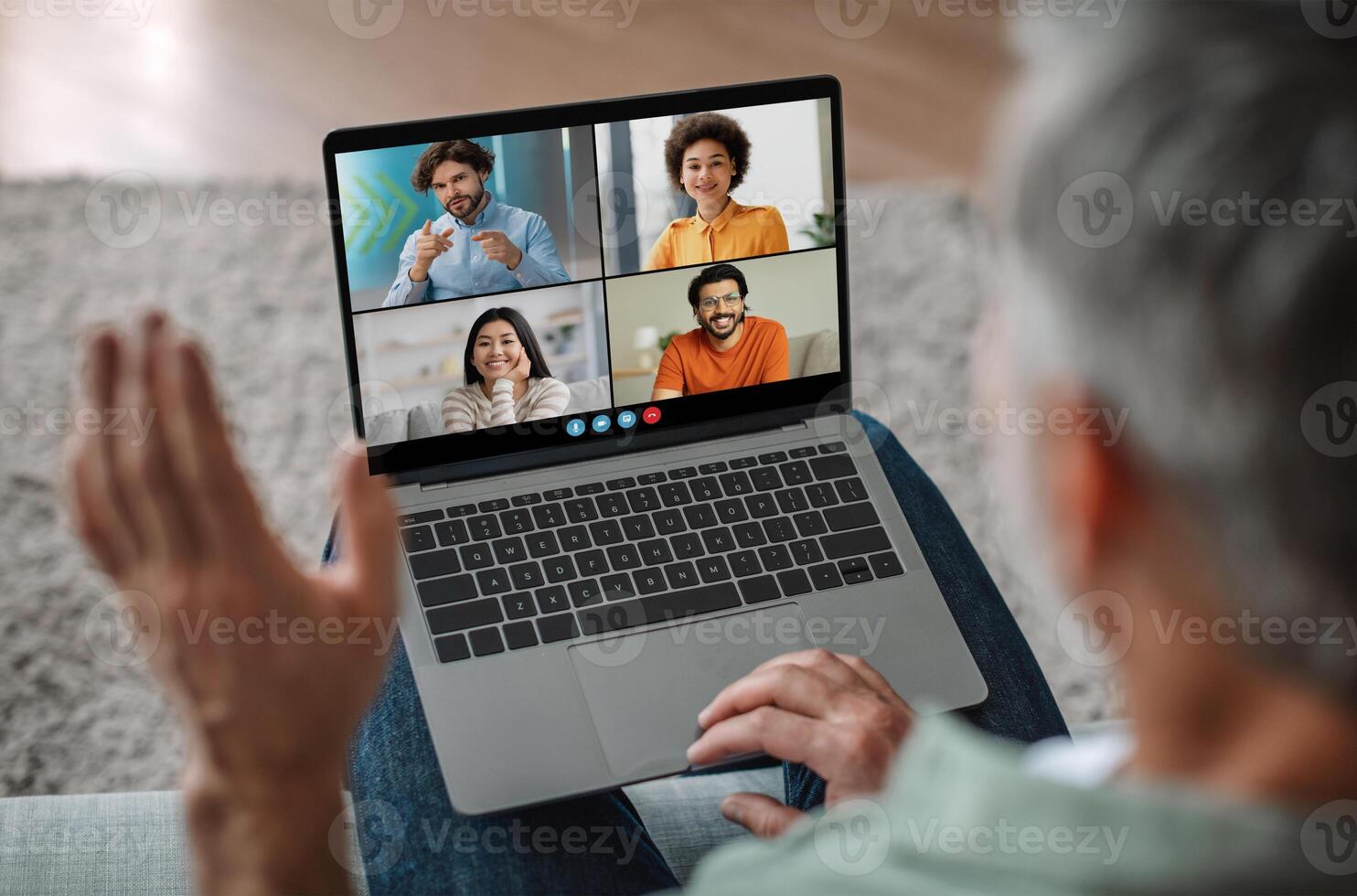 Elderly man sits on a couch, using a laptop to participate in a group chat. The laptop screen shows four tiles of different individuals, each with a different background. photo