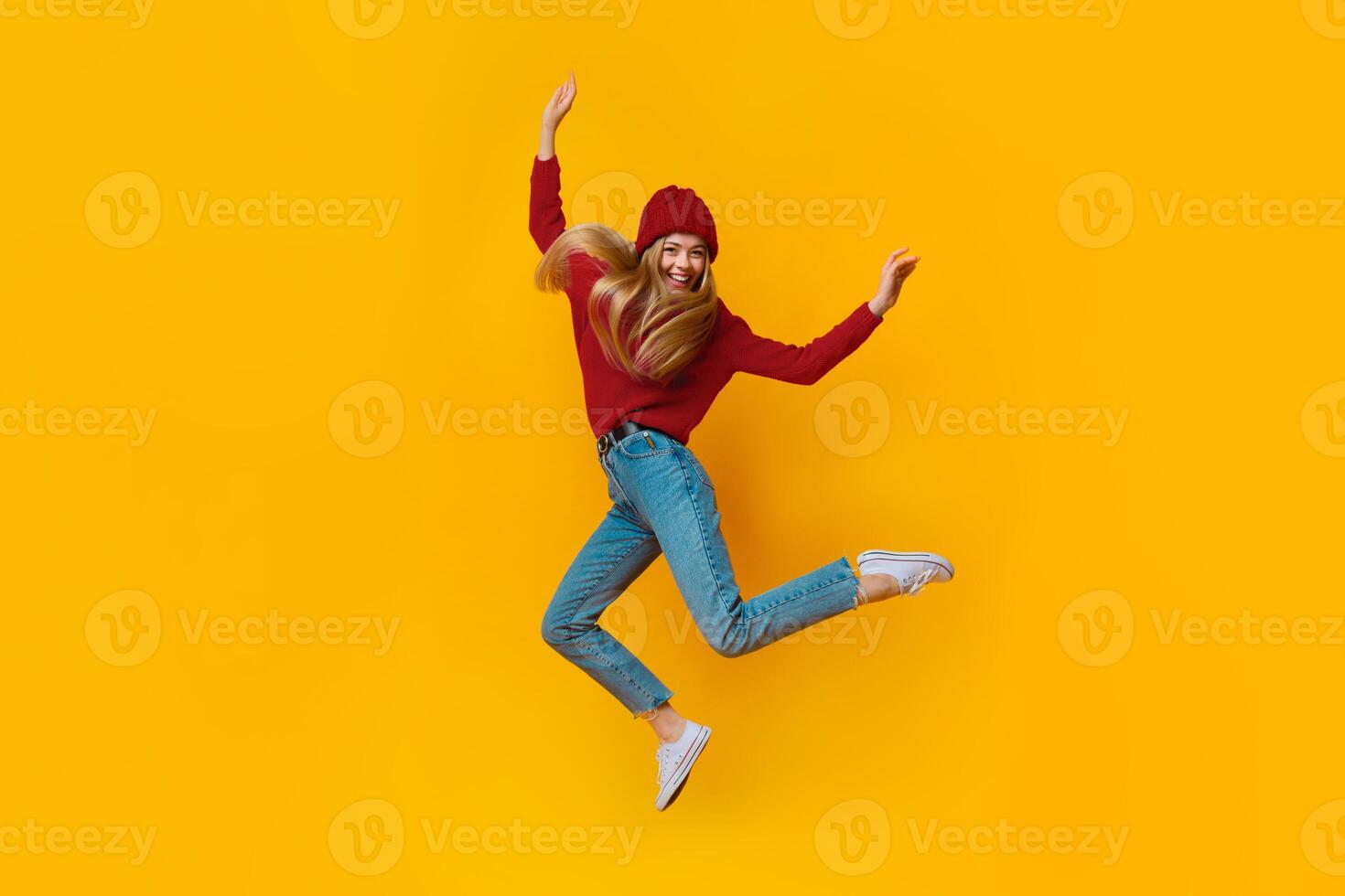 Breezy girl in blue winter clothes jumping in the air, showing funny moves, yellow studio background photo