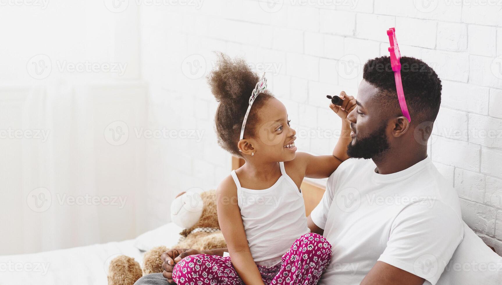African American little girl, wearing a tiara, applies makeup to her fathers face with a brush while sitting on a bed. They are both smiling and having fun together photo