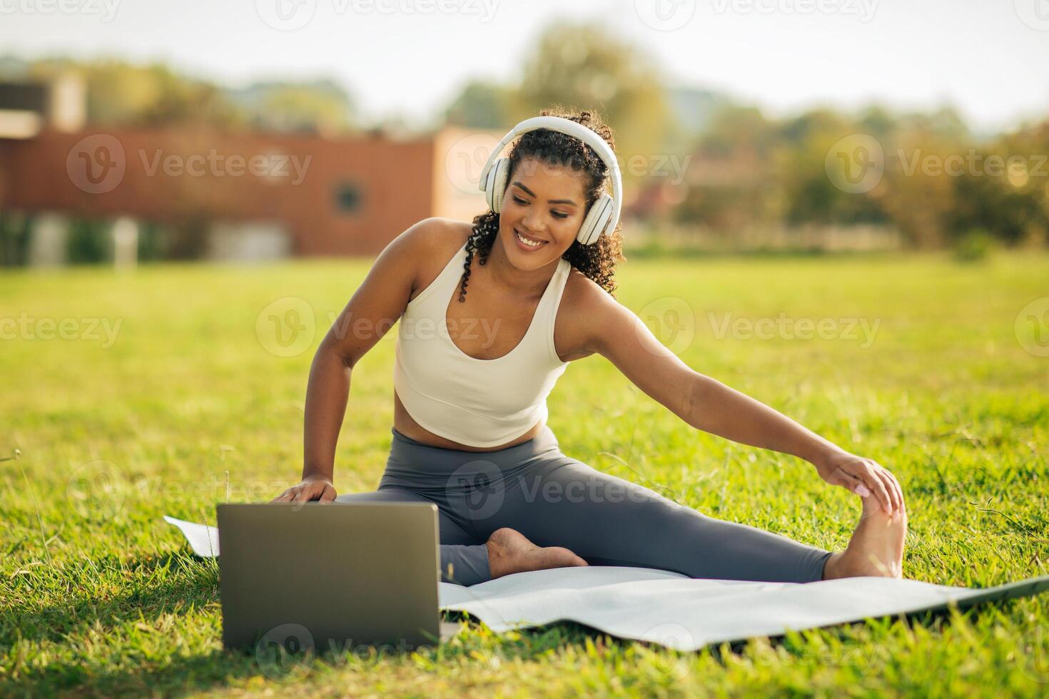 A young woman enjoys a sunny day by practicing yoga on a mat in a grassy field. She wears headphones while stretching her leg, connecting with her mind and body in a serene environment. photo