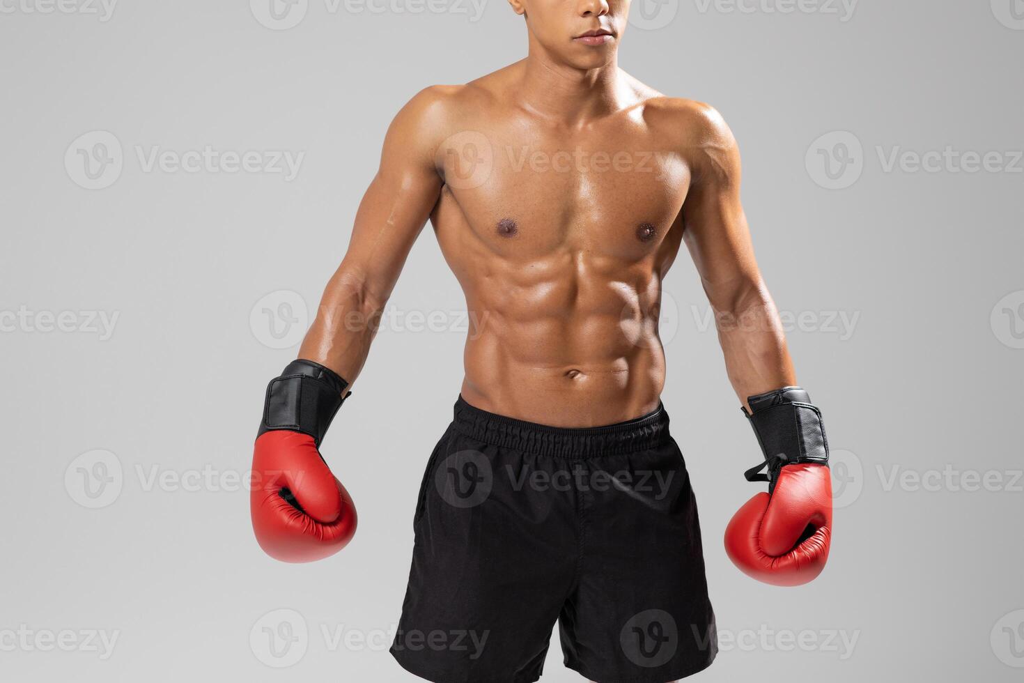 A fit young man boxer stands confidently in a studio, showcasing his defined abs and muscular arms while wearing vibrant red boxing gloves, cropped photo