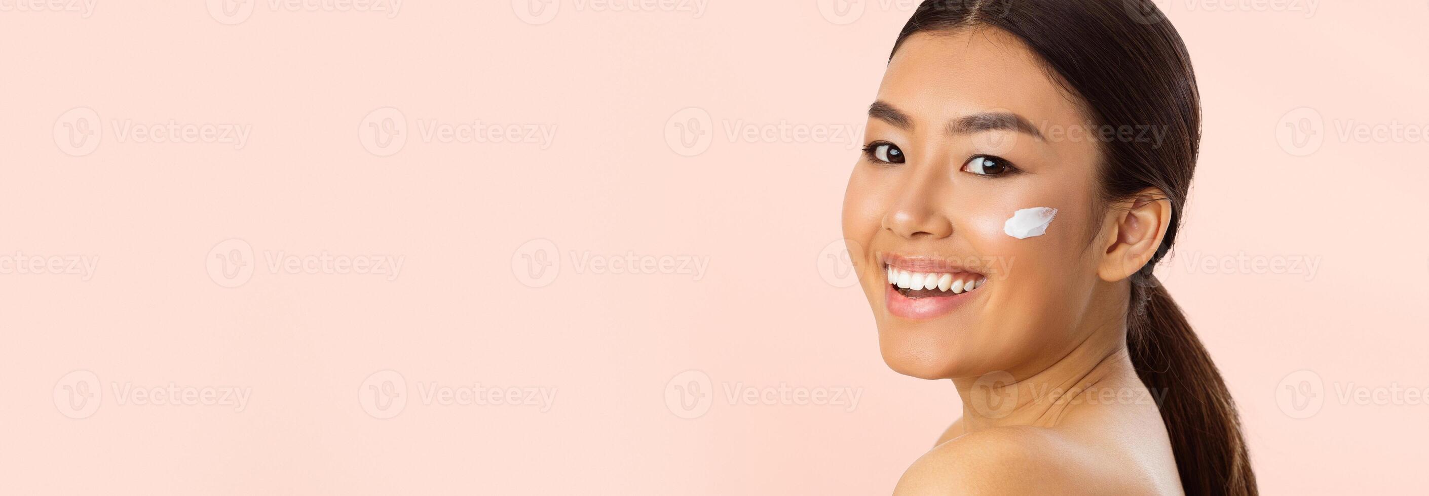 Asian woman with dark hair and brown eyes smiling as she applies white moisturizer to her cheek. She has a natural look and is wearing a low ponytail. The background is a soft pink color, copy space photo