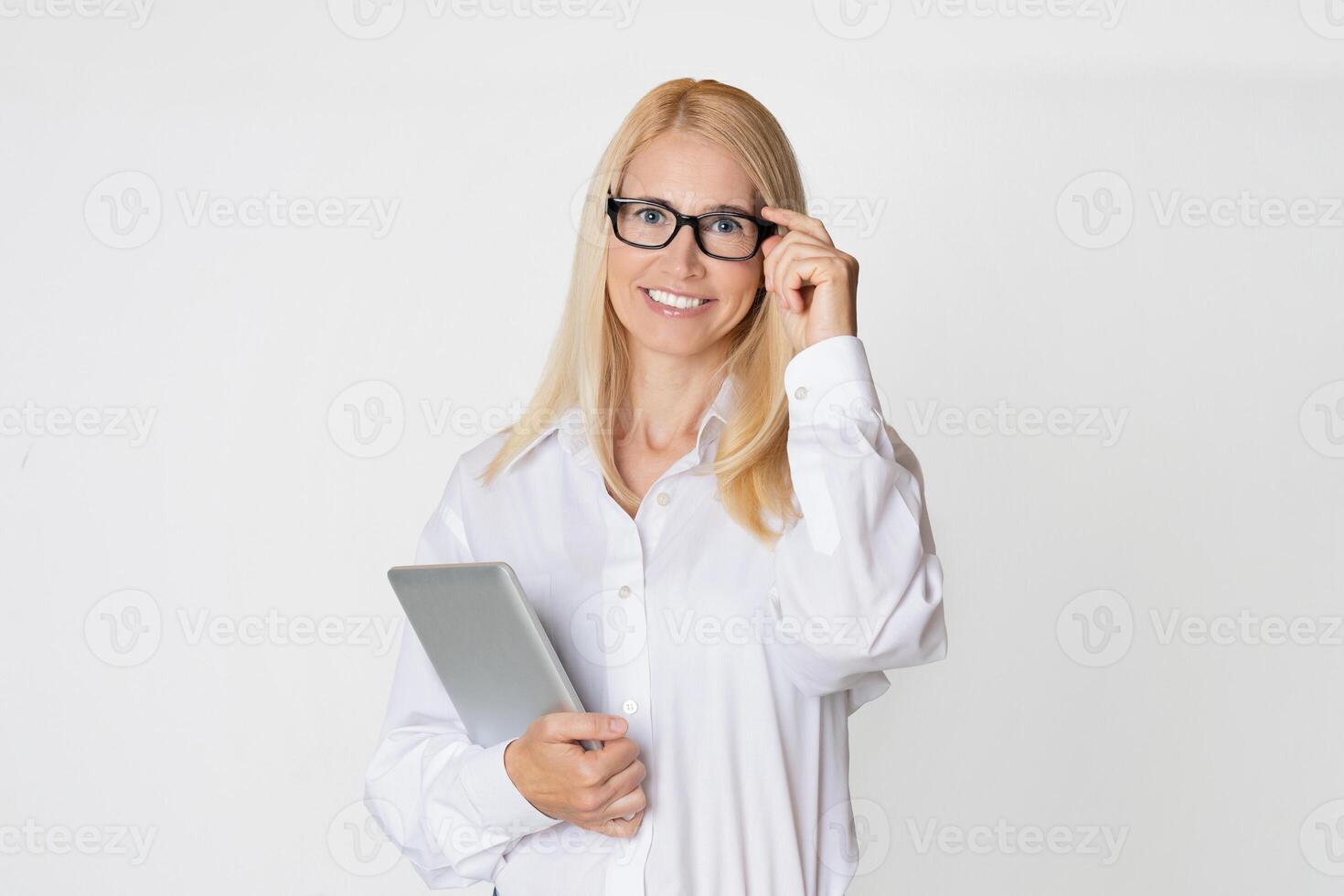 A young, attractive businesswoman wearing glasses smiles and looks directly at the camera. She is holding a tablet computer in her left hand and adjusting her glasses with her right hand. photo