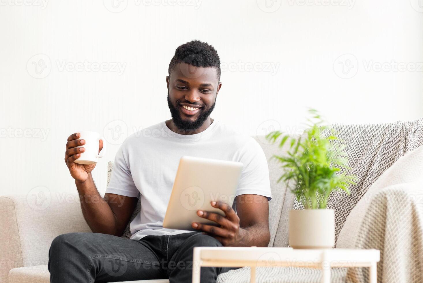 A happy African American man sits on a couch in a living room, holding a tablet in one hand and a mug of coffee in the other. He is smiling and looks relaxed. photo