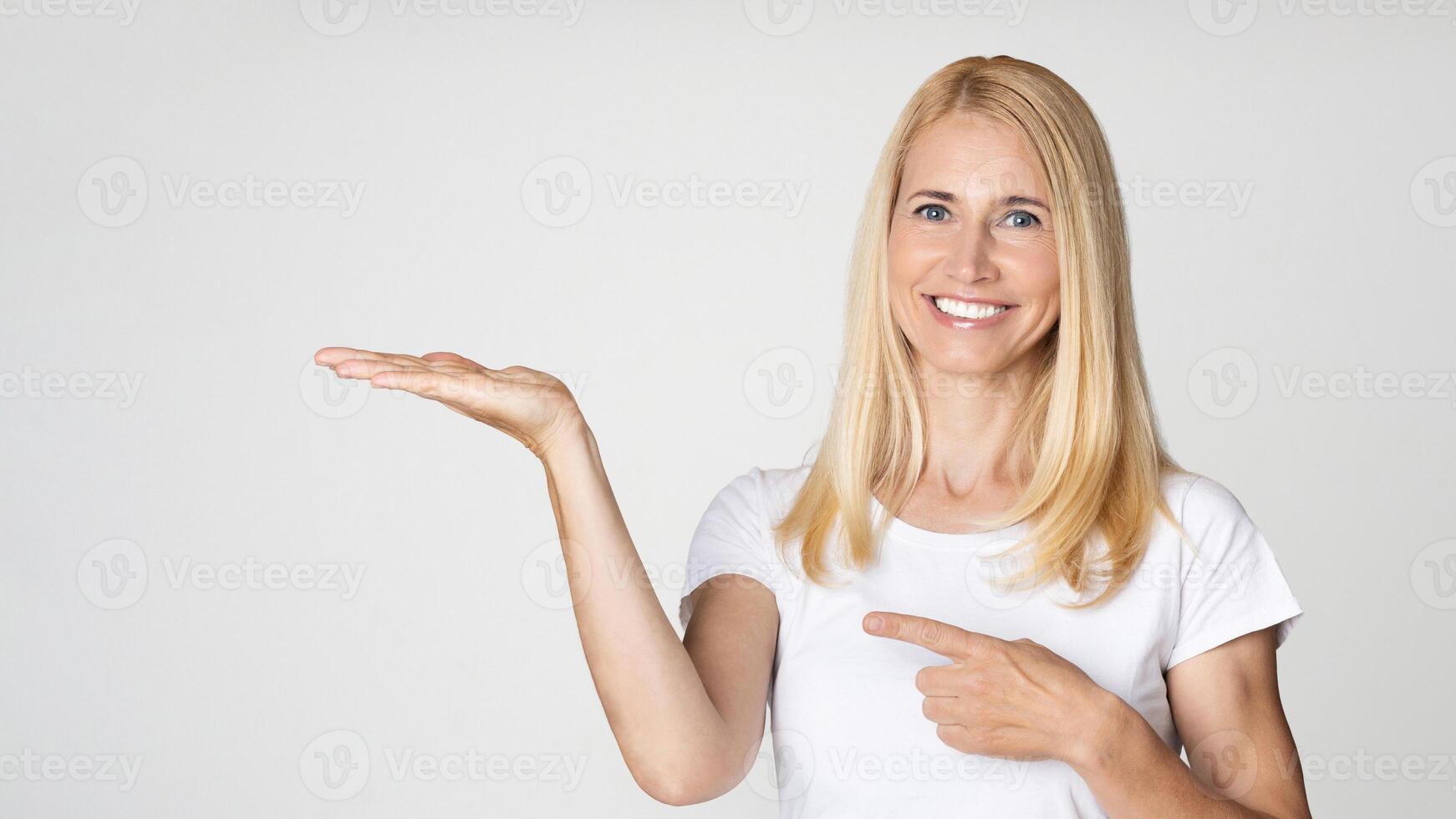A happy blonde woman smiles and points to her open hand, presenting a product against a white background. Her expression is warm and inviting, suggesting she is excited about the product photo