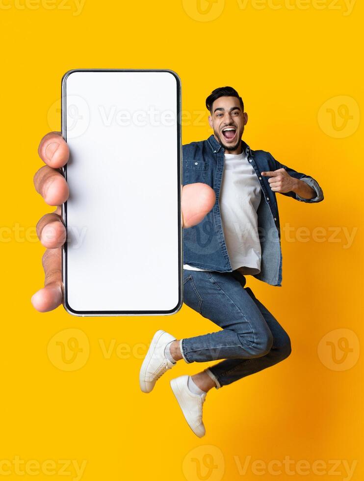 Handsome young Arab guy jumping on air, pointing at cellphone with empty screen on orange studio background. Millennial Muslim man demonstrating mockup for mobile app design photo