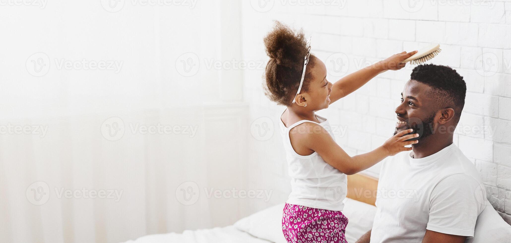Black little girl with dark curly hair, wearing a white tank top and floral shorts, is brushing her fathers hair with a wooden brush. Man is smiling and looking at his daughter as she brushes his hair photo