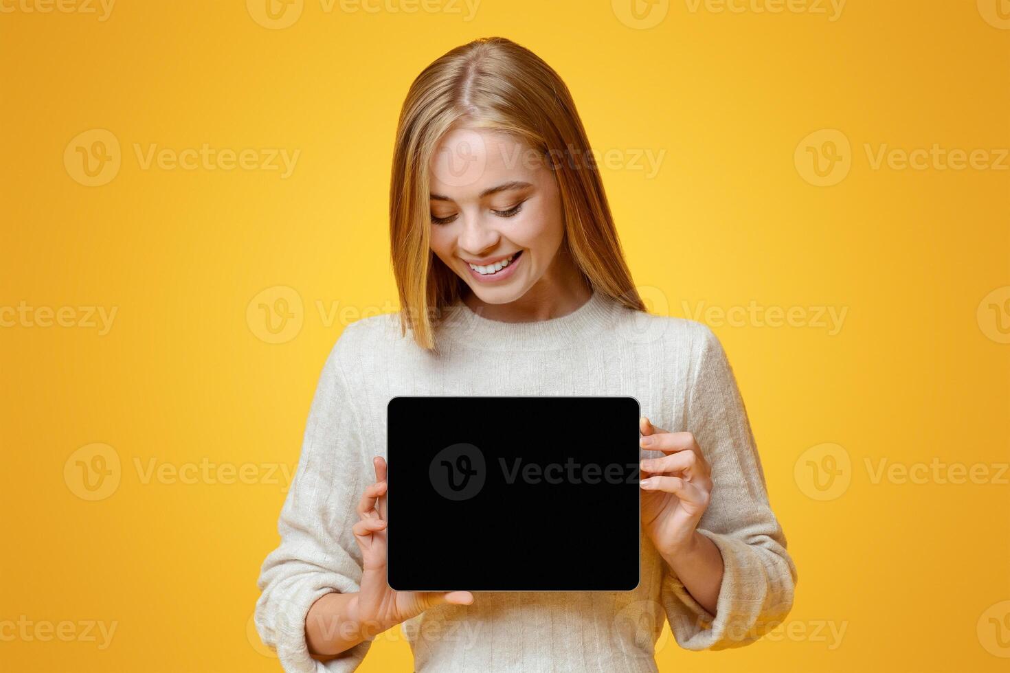 Smiling millennial girl looking at digital tablet with blank screen in her hands, orange studio background with copy space photo
