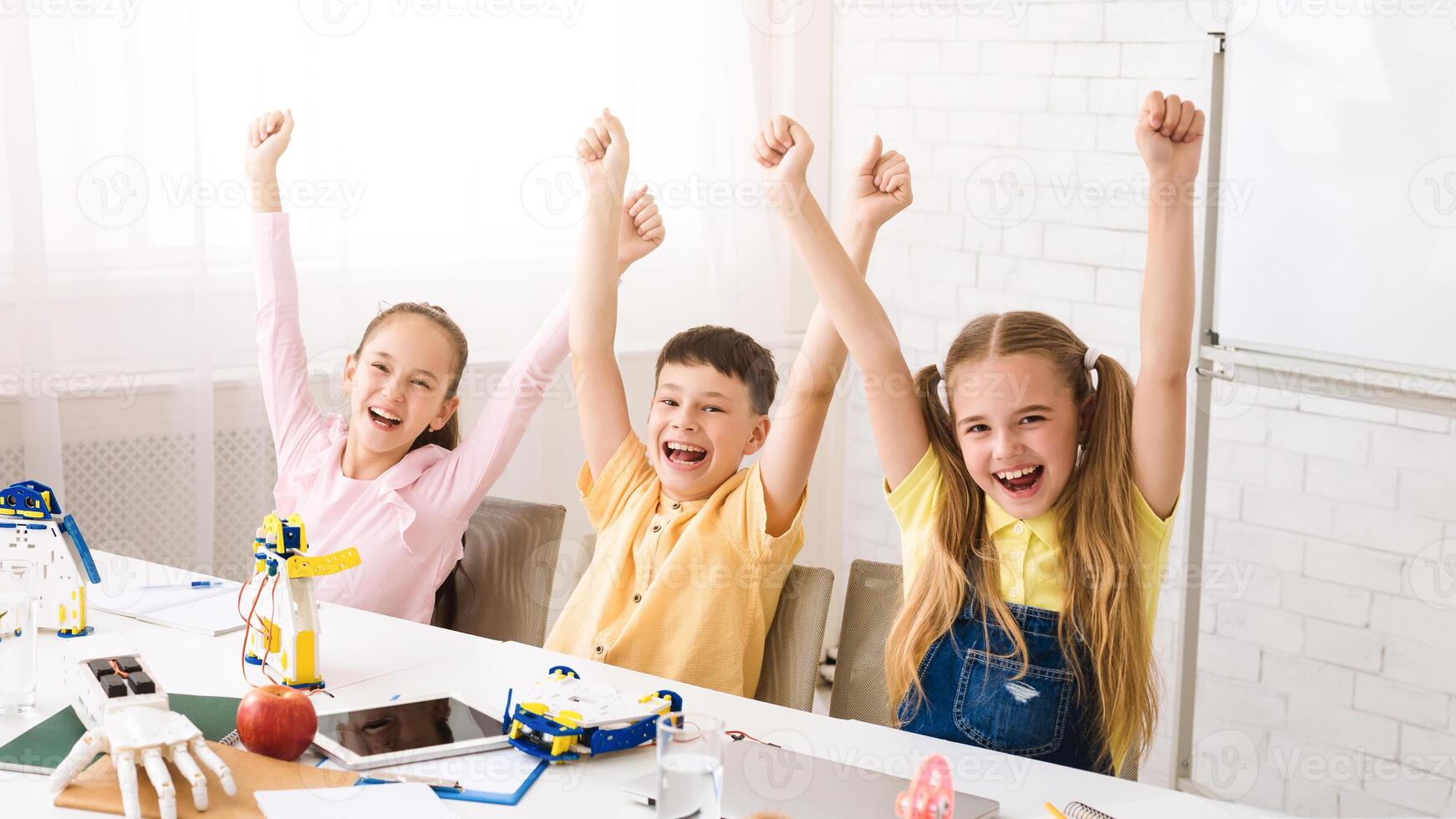 Three children, two girls and one boy, sit at a table with their arms raised in victory. They have completed a successful STEM project and are beaming with pride. photo