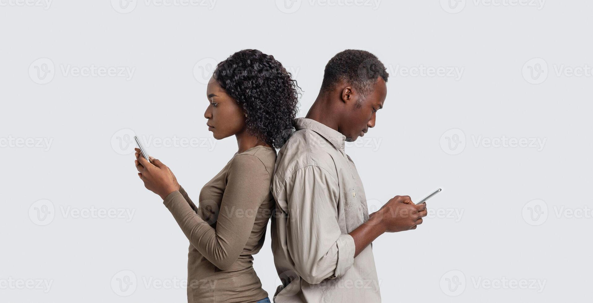 African american man and woman standing back to back, using mobile phones during quarantine, grey studio background, copy space. Black couple sitting in social media instead of talking to each other photo