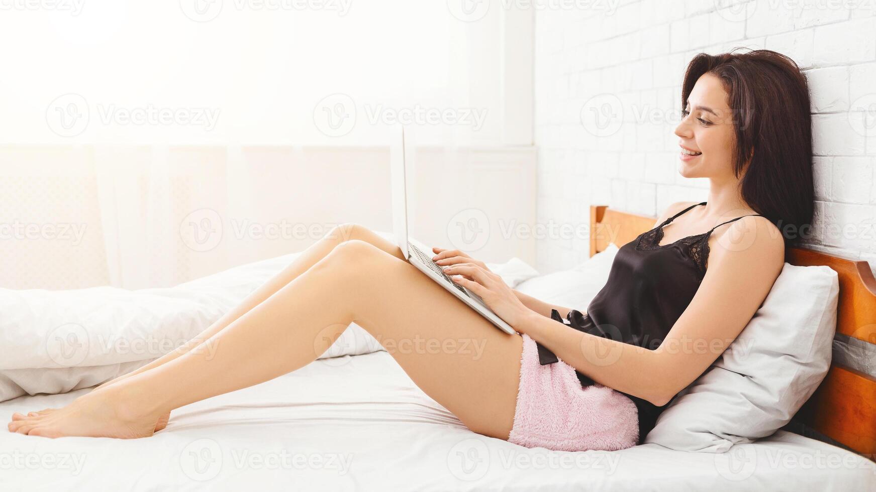 A woman is sitting comfortably on a bed, focused on using a laptop computer. She appears engaged in her work or leisure activity, with the screen reflecting in her eyes, side view photo