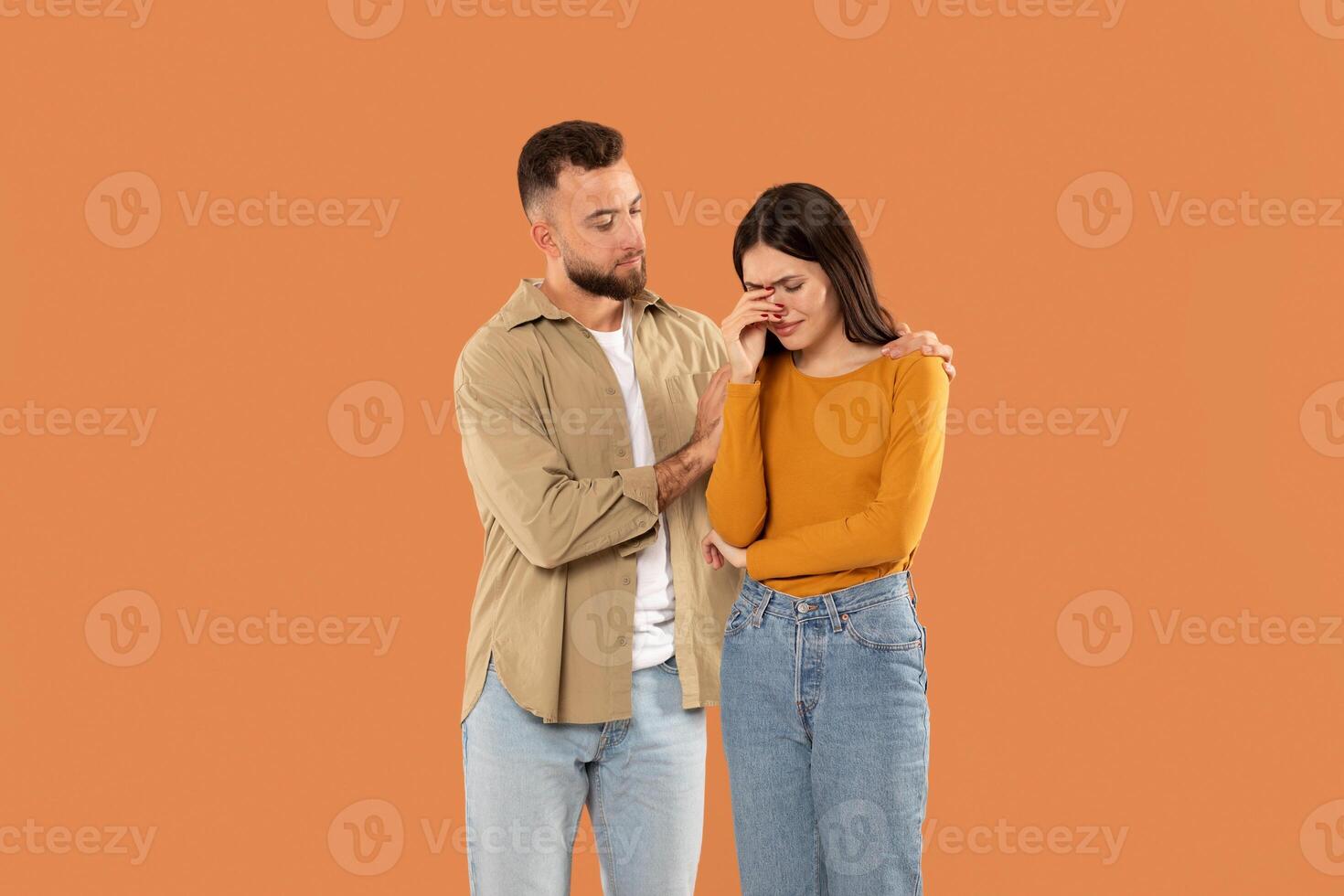 A man stands next to a distressed woman, offering her comfort and support. He places a reassuring hand on her shoulder while she wipes tears from her eyes. They are dressed in casual photo