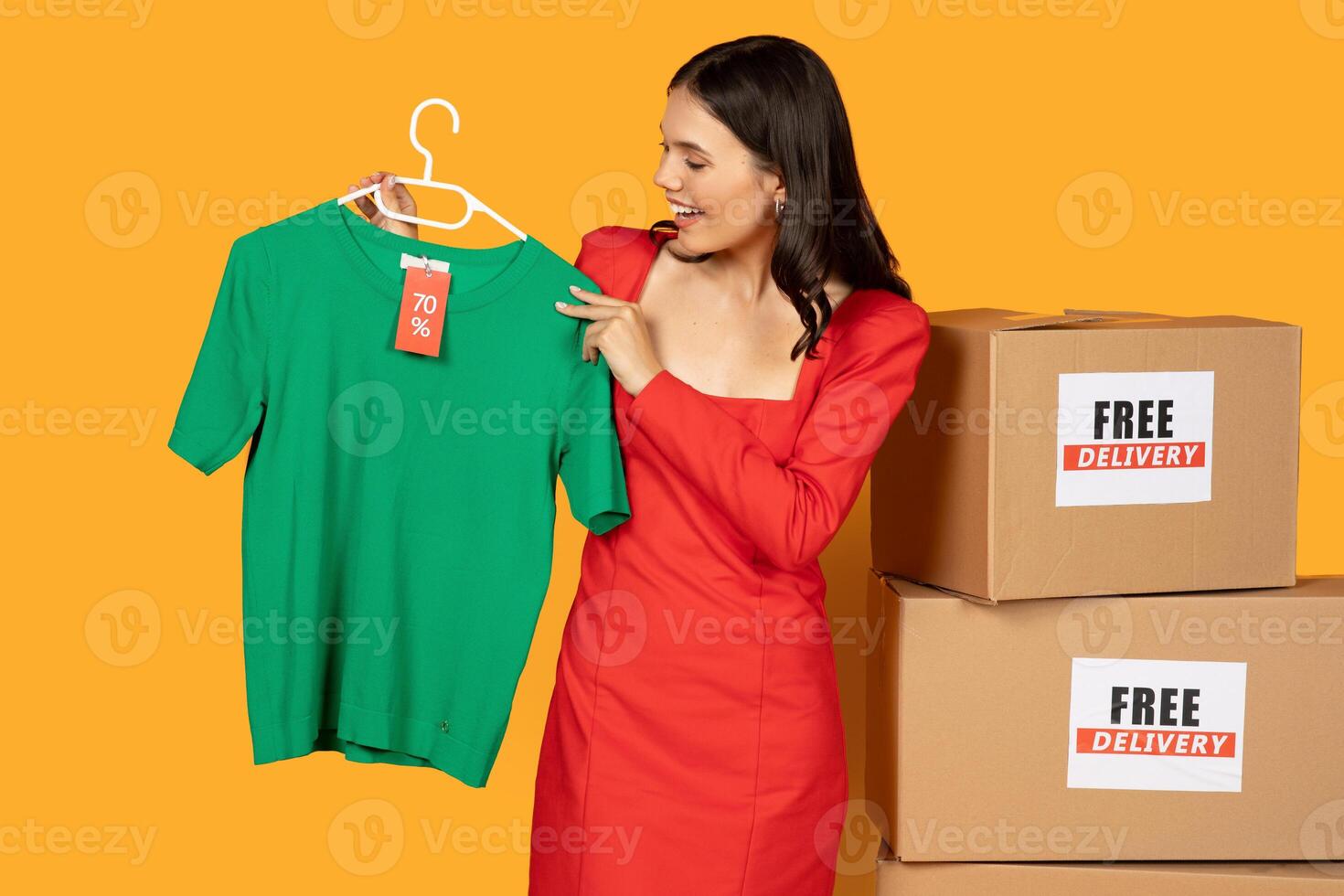 A woman in a red dress stands in front of a bright yellow background holding a green t-shirt with a 70 off tag on a hanger. Behind her are two stacked cardboard boxes labeled Free Delivery. photo