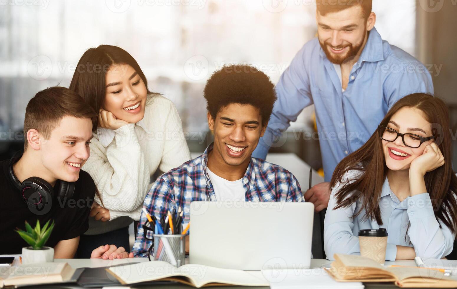 A vibrant group of four young adults are gathered around a table filled with books, engaging in a cheerful conversation with focus on a laptop screen, doing homework together photo