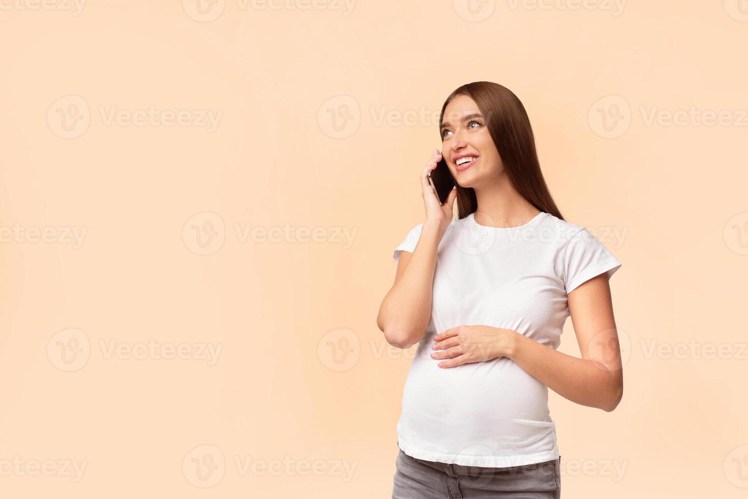 Cheerful Pregnant Girl Talking On Phone Consulting With Doctor Standing In Studio Over White Background. Empty Space photo