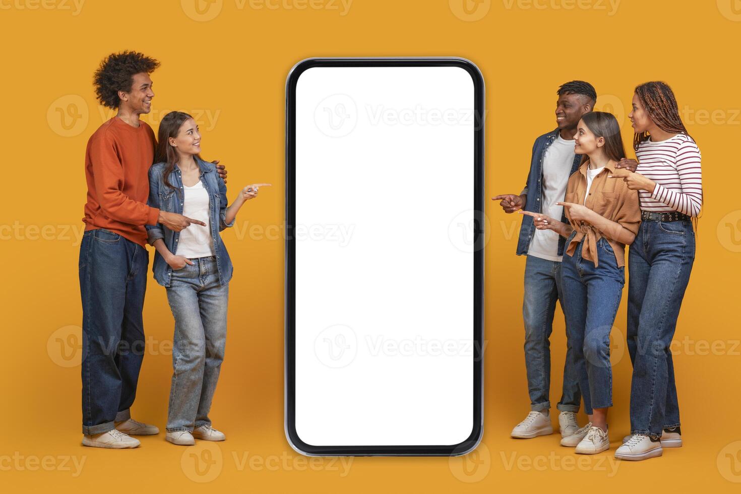 A group of five friends stand in front of a large smartphone with a blank white screen. They are all pointing at the screen, suggesting they are interested in what is being displayed photo