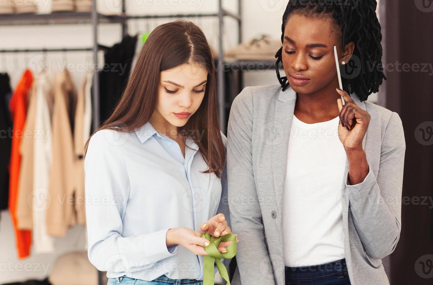 Two multiethnic women stand side by side in clothing store, carefully examining the material of a garment. The thoughtful expression on their faces suggests they are making a considered choice photo