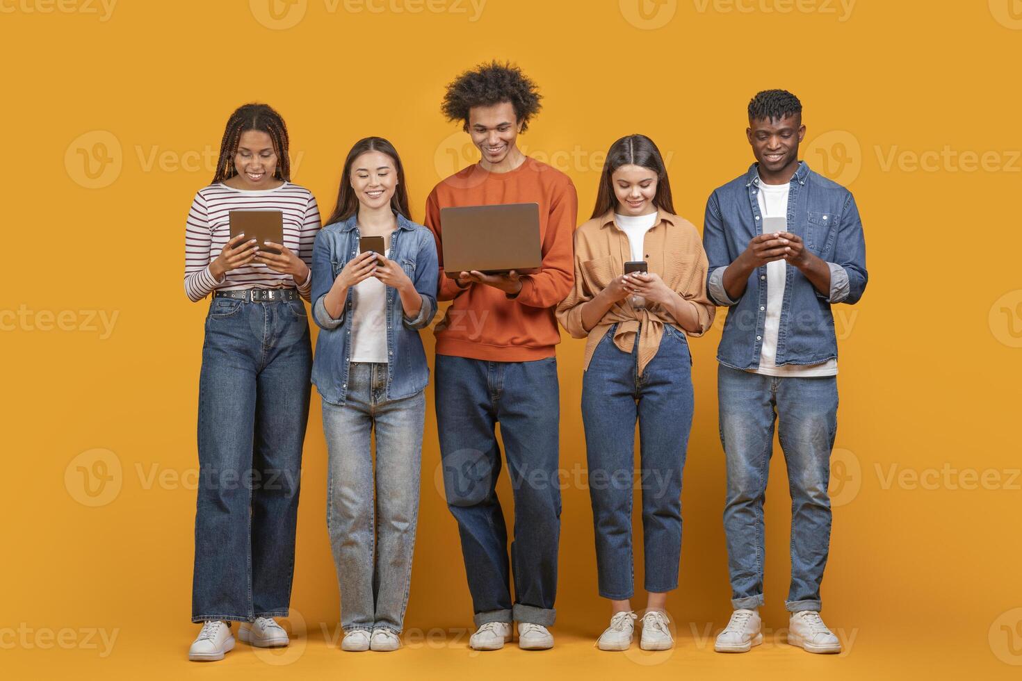 This image shows a group of five young adults standing in a row against a yellow background. They are all looking down at their devices, which include a tablet, a smartphone, and a laptop photo