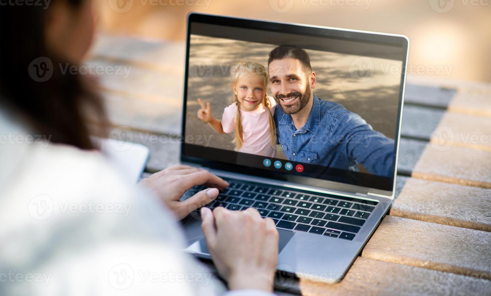 A woman sits at a wooden table outdoors and uses a laptop to chat with her family. The screen displays a man and his daughter, who are both smiling and waving. photo
