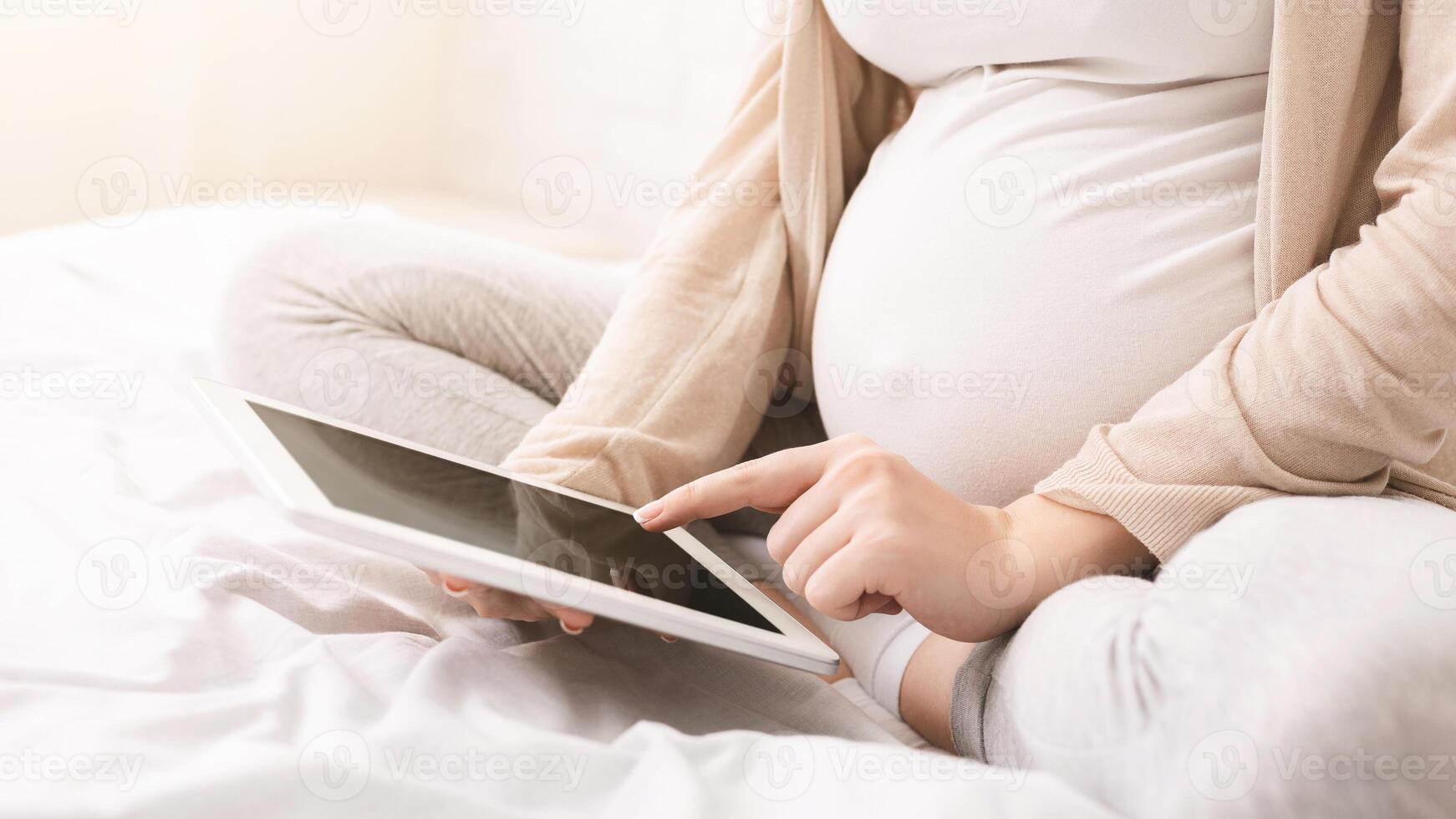 A pregnant woman is sitting on a bed, holding a tablet with black screen in her hands, possibly reading or watching something. The room is simple and comfortable, cropped photo