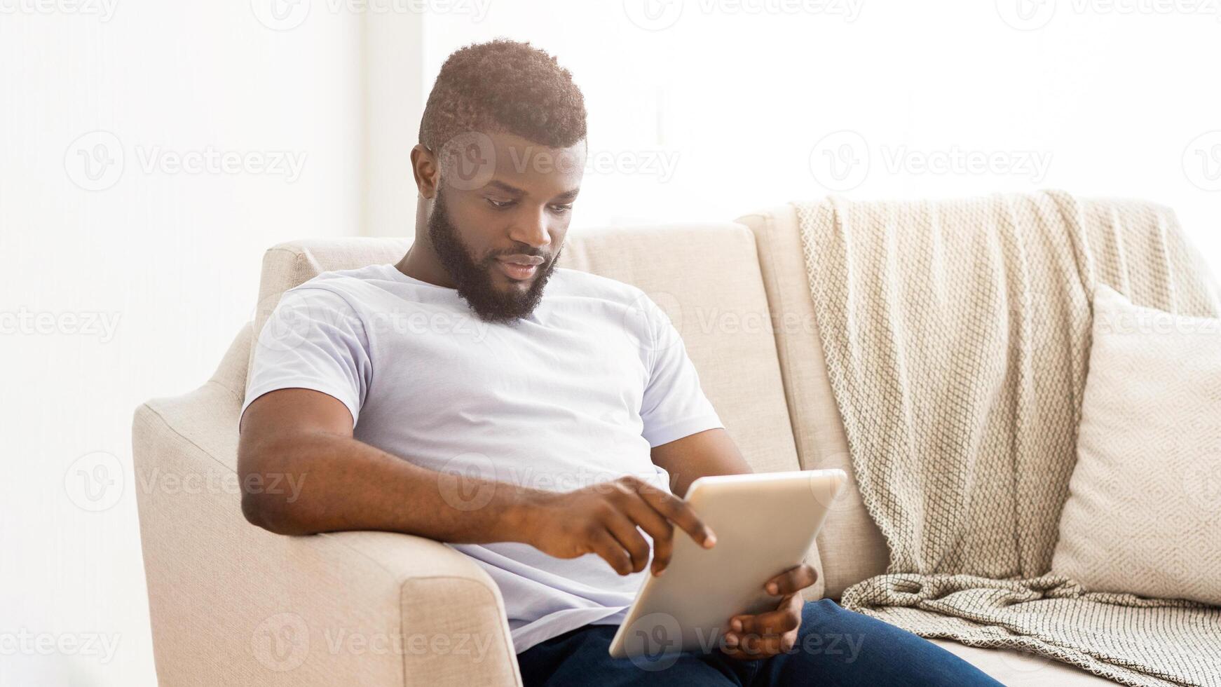 African American man is sitting on a couch in a living room. He is relaxed and using a tablet device. The room is brightly lit, and the man appears to be focused on the screen. photo